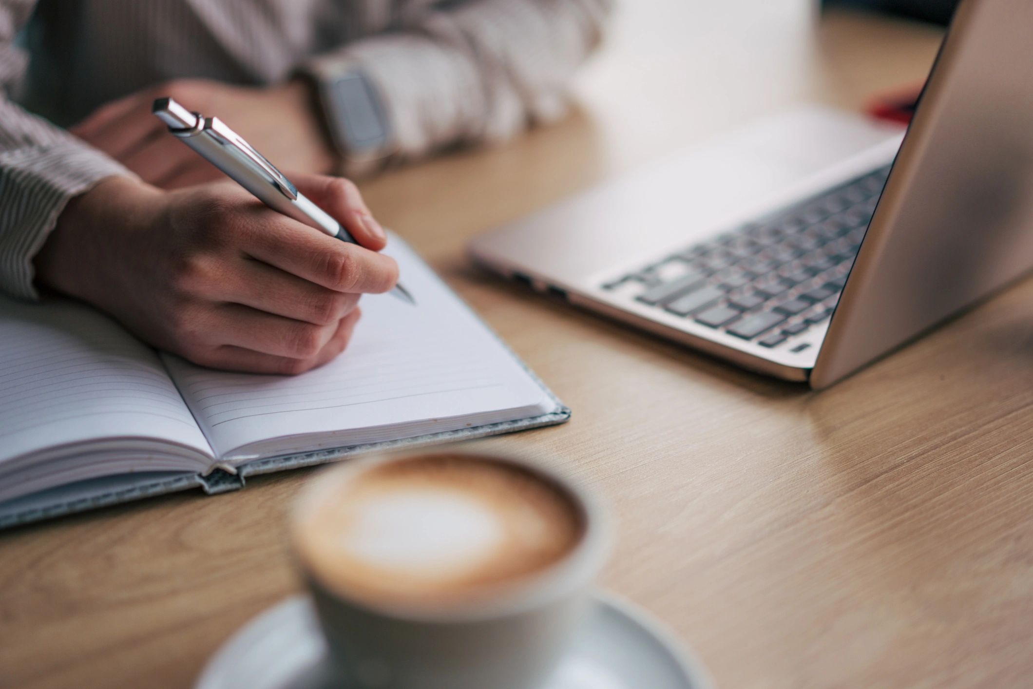 Hands writing in a notebook beside a laptop in natural light at a home desk